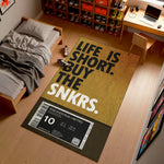 Doormat with 'Life is short, buy the snkrs' text in a room with a bed and shelves.

