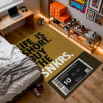 Doormat with 'Life is short, buy the sneakers' text in a room with shoes and a laptop.


