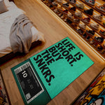 Green doormat with text on a wooden floor next to a bed and shoe rack.

