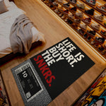 Doormat with 'Life is short, buy the snkers' text on a wooden floor next to a bed.

