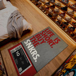 Doormat with 'Life is short, buy the SNKRS' text on a wooden floor next to a bed and shoe rack.

